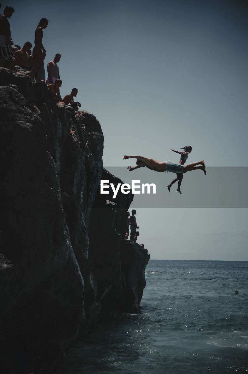 LOW ANGLE VIEW OF PEOPLE JUMPING ON ROCK FORMATION IN SEA