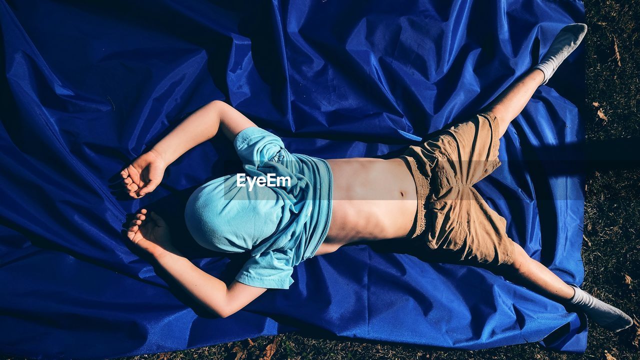 Full length of boy with covered face sleeping on blue fabric in yard