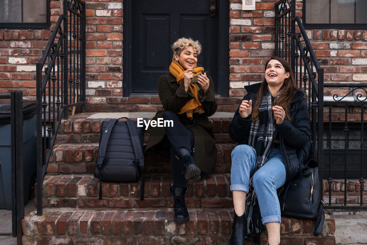 Happy female friends talking while sitting on steps in city during winter