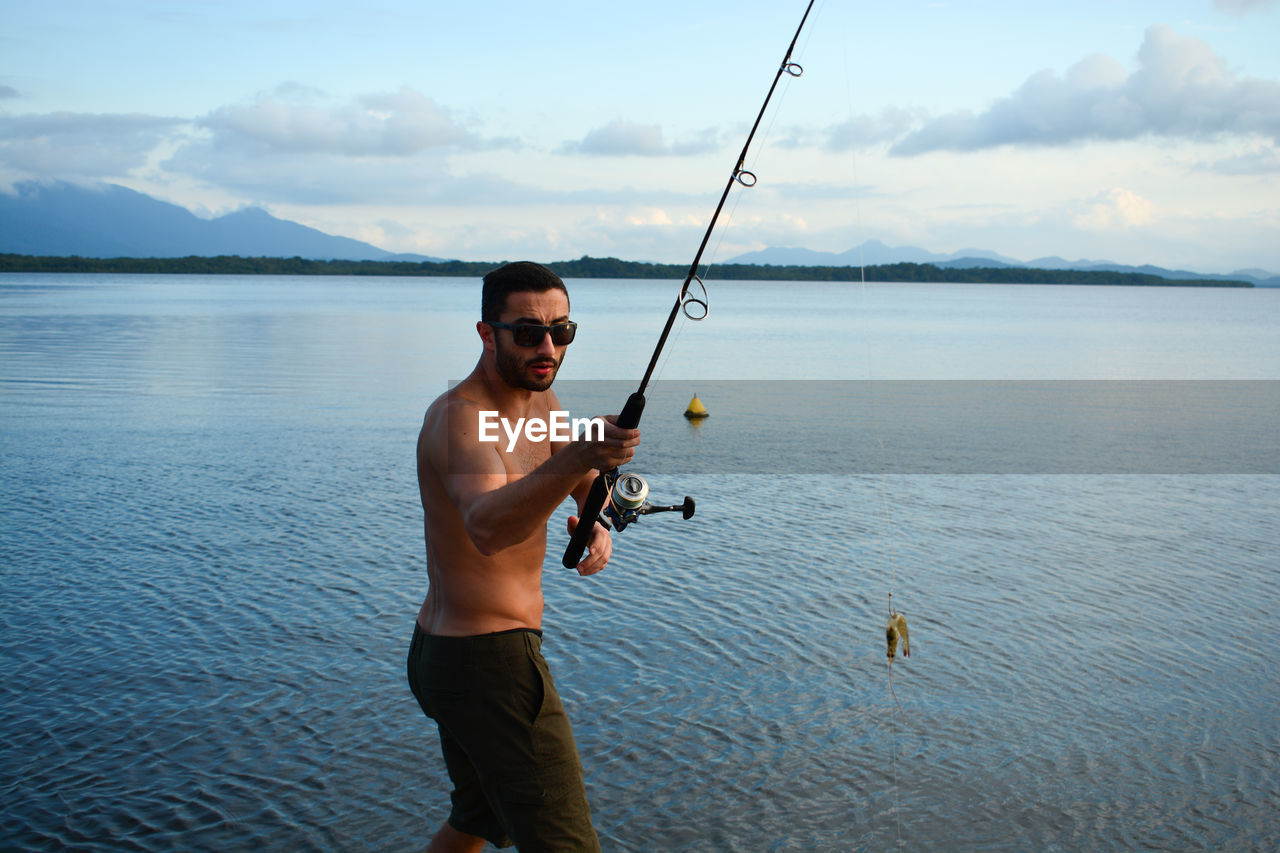 Portrait of young man holding fishing rod while standing in lake against sky