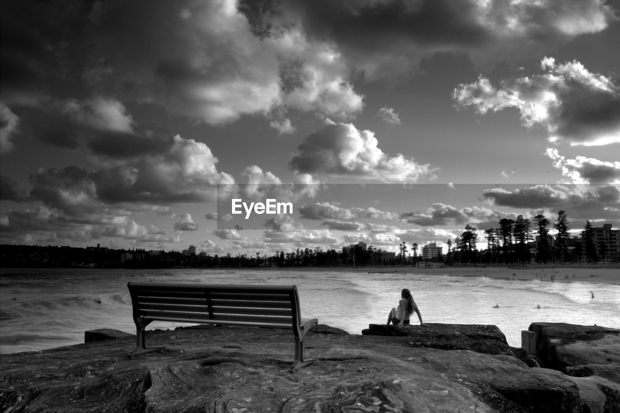 Woman sitting at manly beach against cloudy sky at dusk