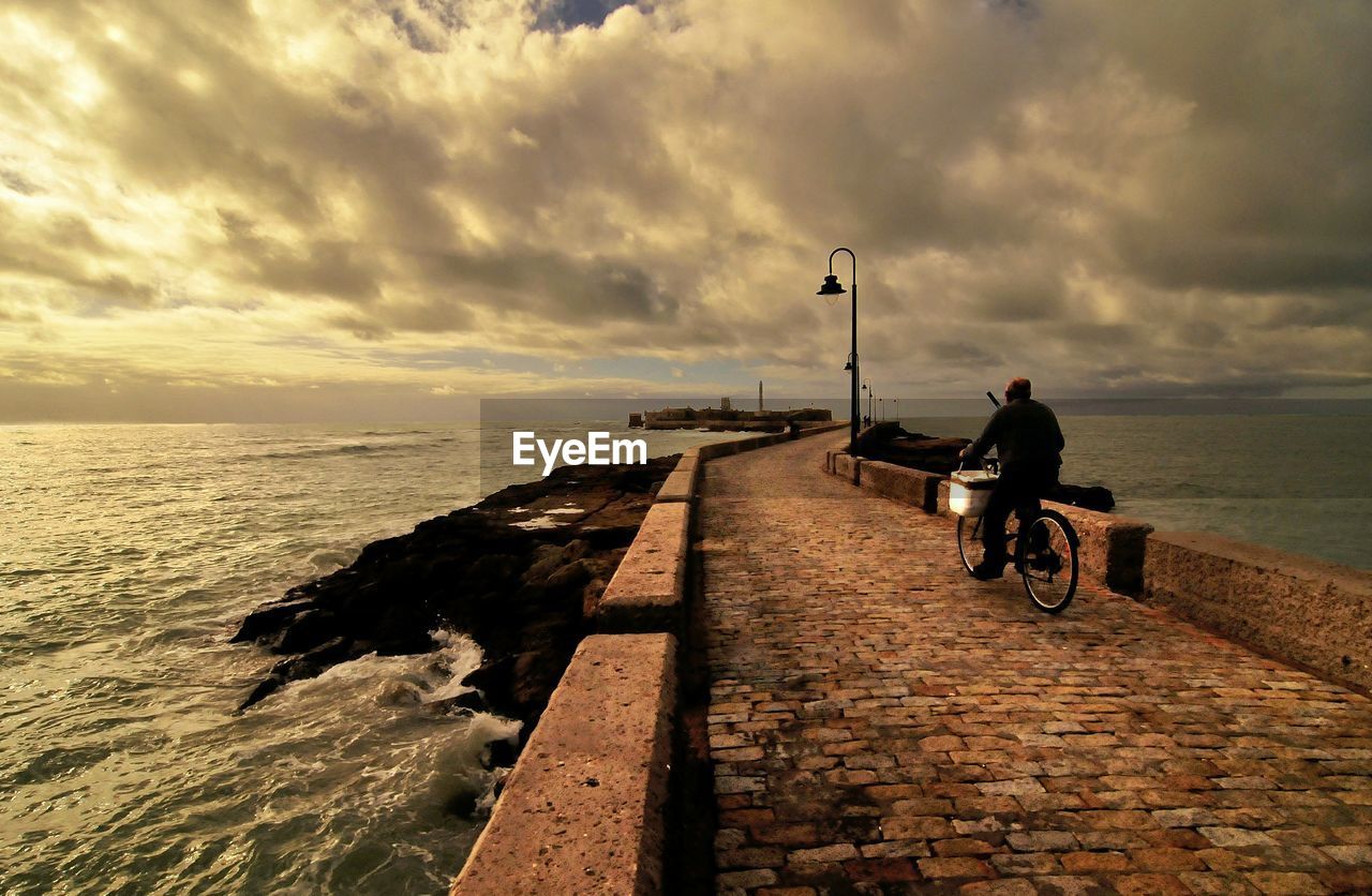 Man riding bicycle on pier against sky during sunset