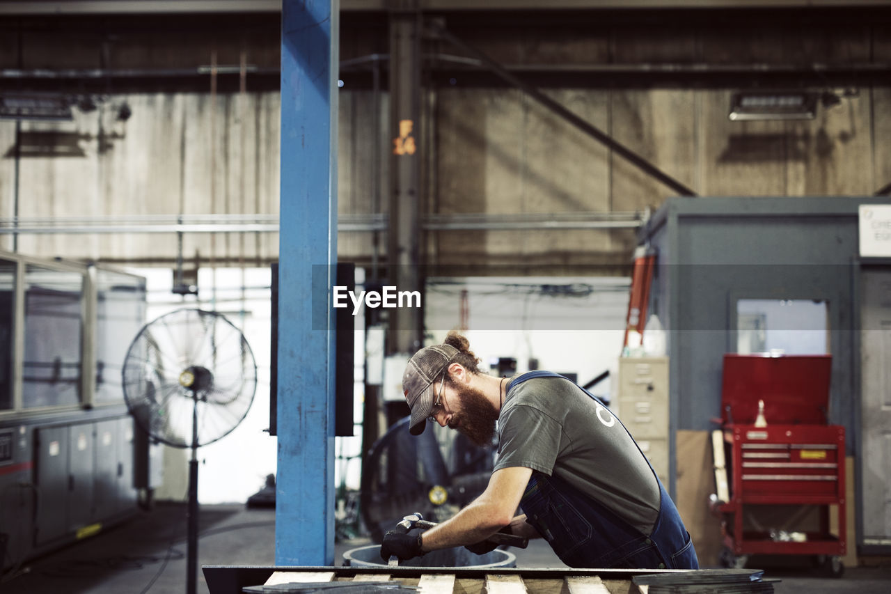 Side view of blue collar worker using hammer and chisel in steel factory