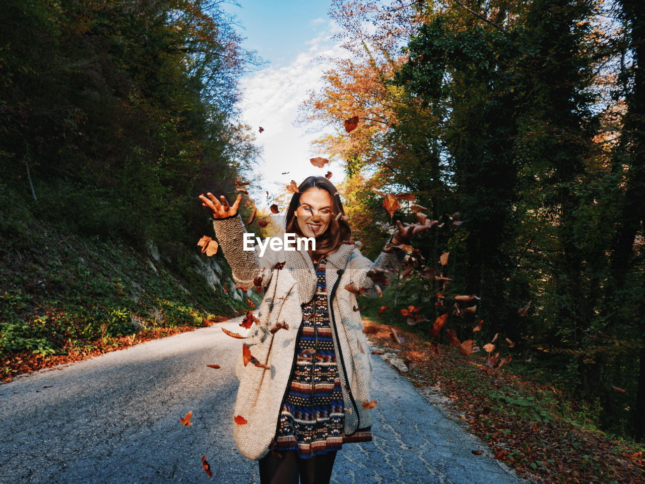 Smiling woman throwing leaves while standing on road against trees