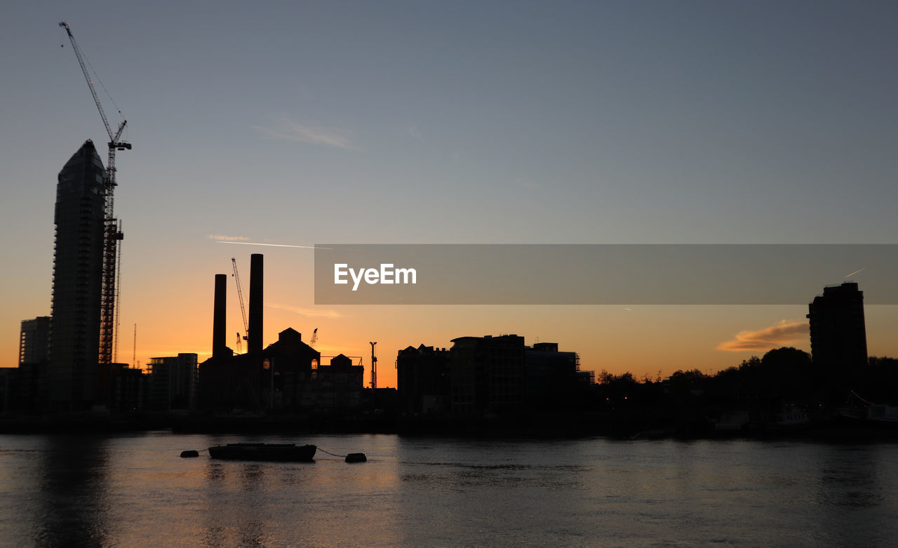 Silhouette buildings by river against sky during sunset