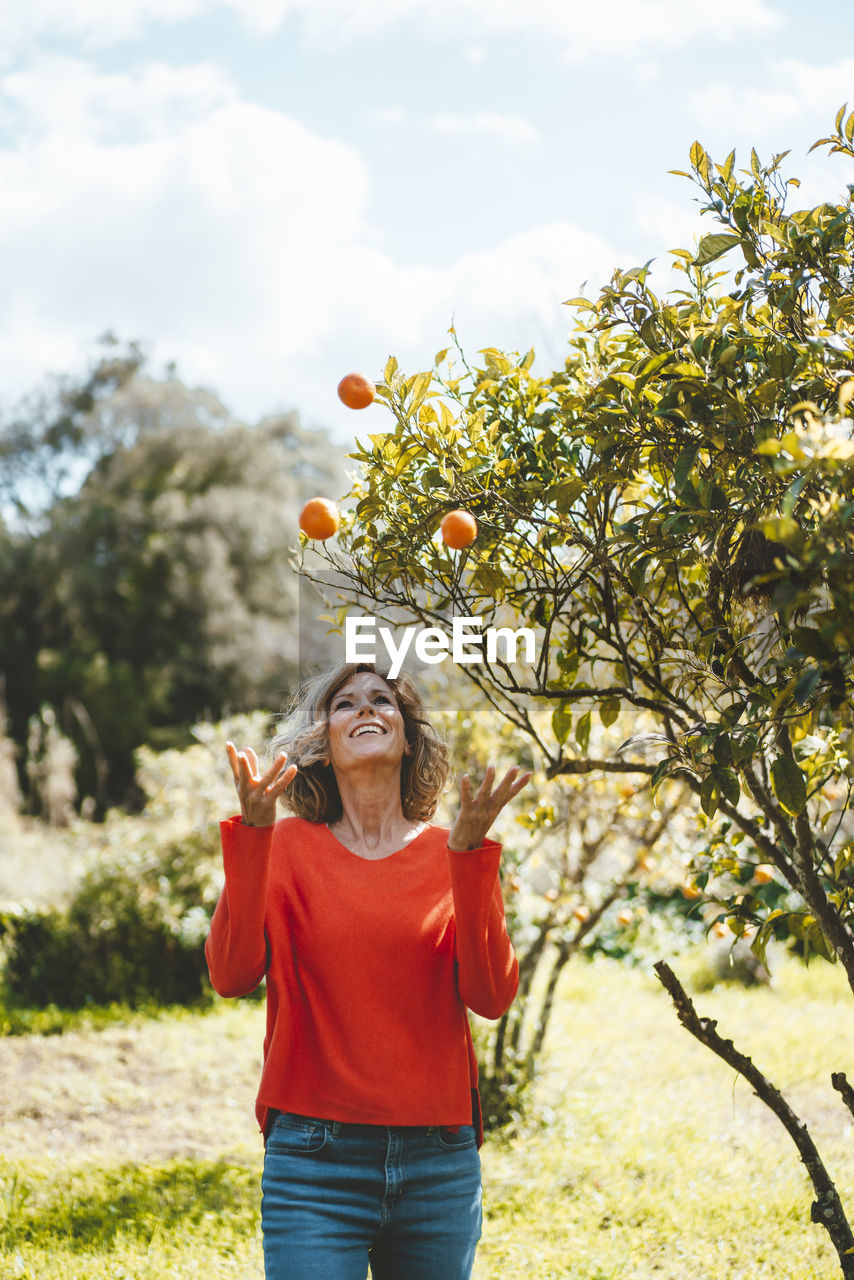 Happy woman juggling oranges standing by tree in garden