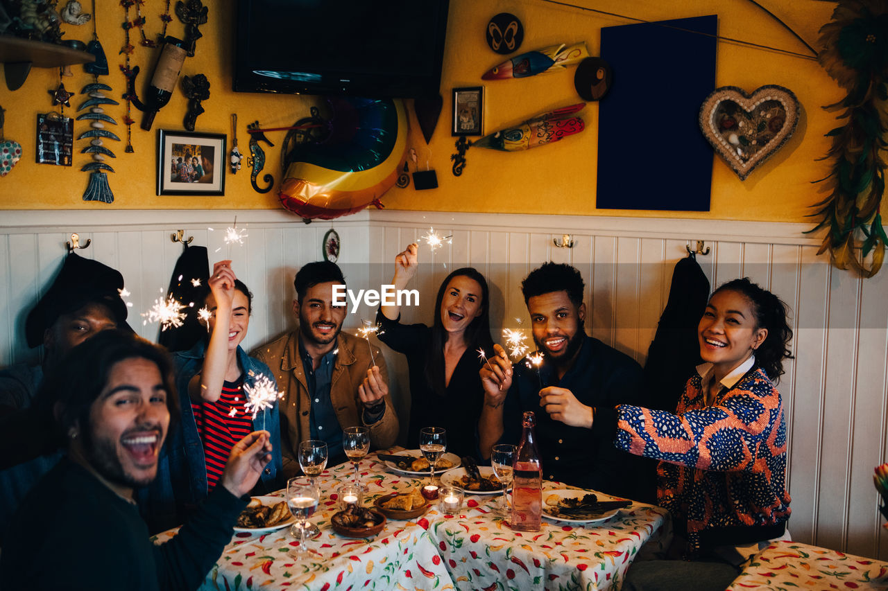 Portrait of cheerful young multi-ethnic friends sitting with sparklers at table in restaurant
