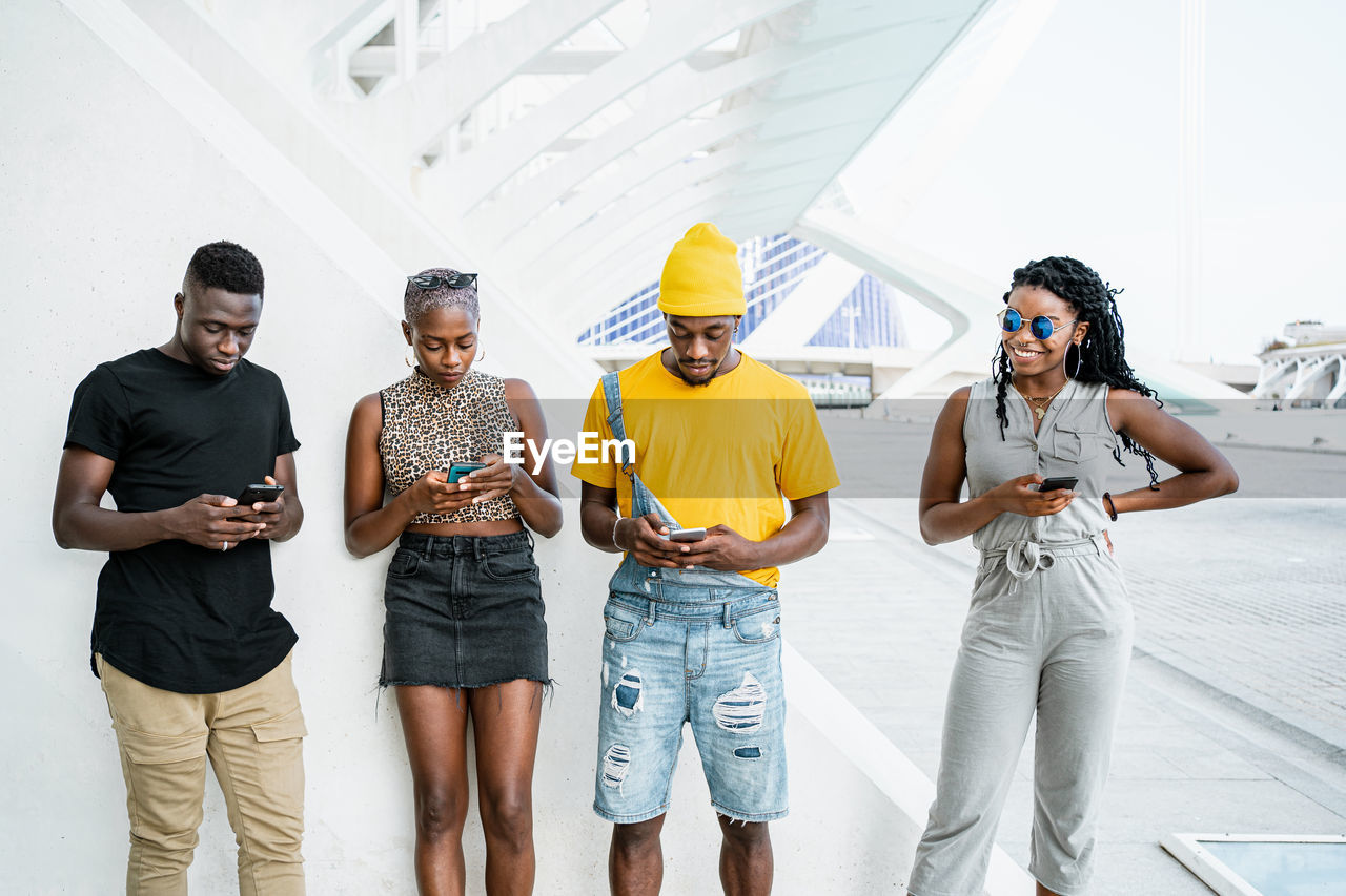 Young black people in stylish clothes standing in urban area and messaging on social media via smartphones