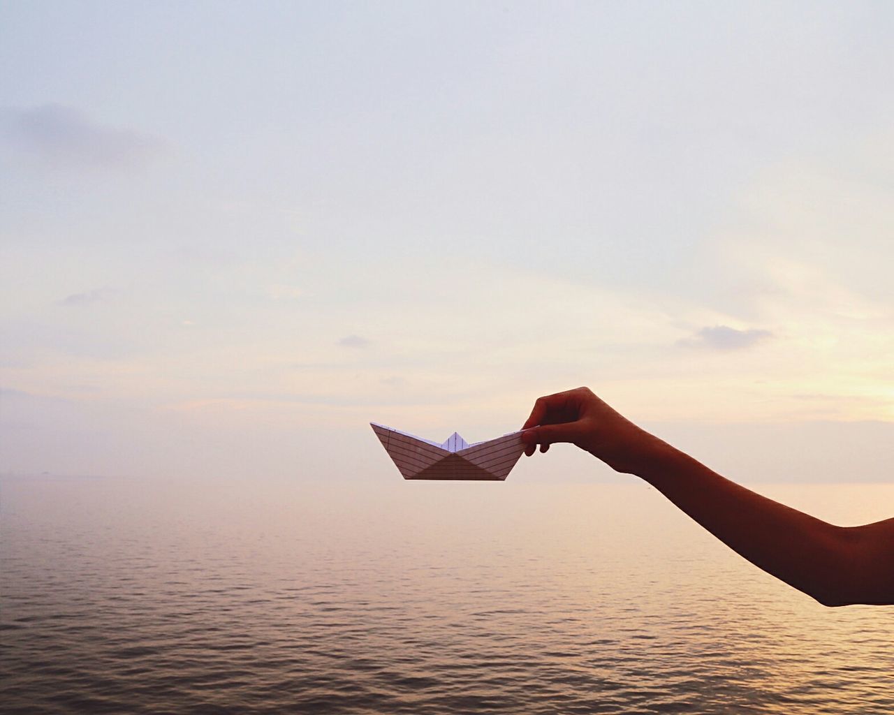 Cropped image of hand holding paper boat over sea against sky