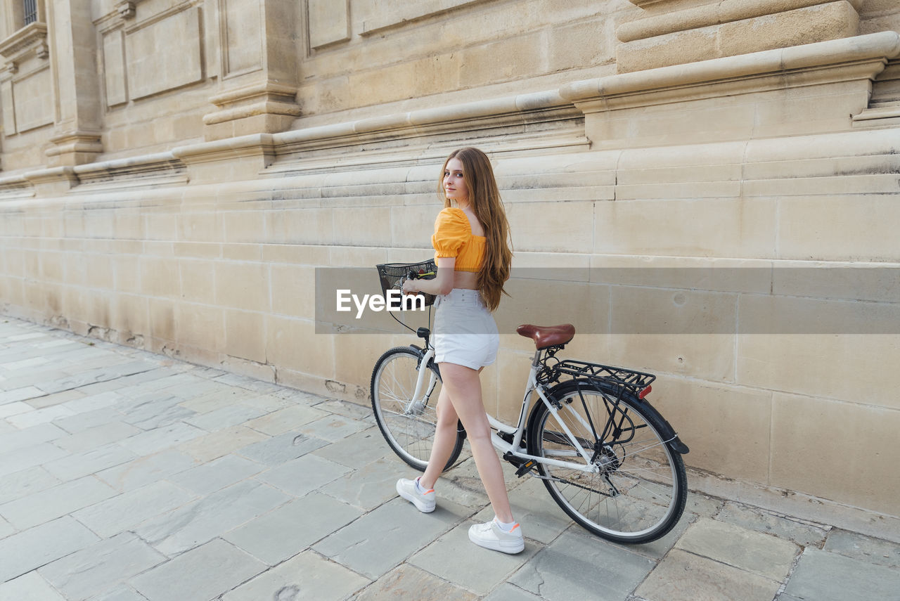 Young woman looking away while wheeling bicycle by stone wall