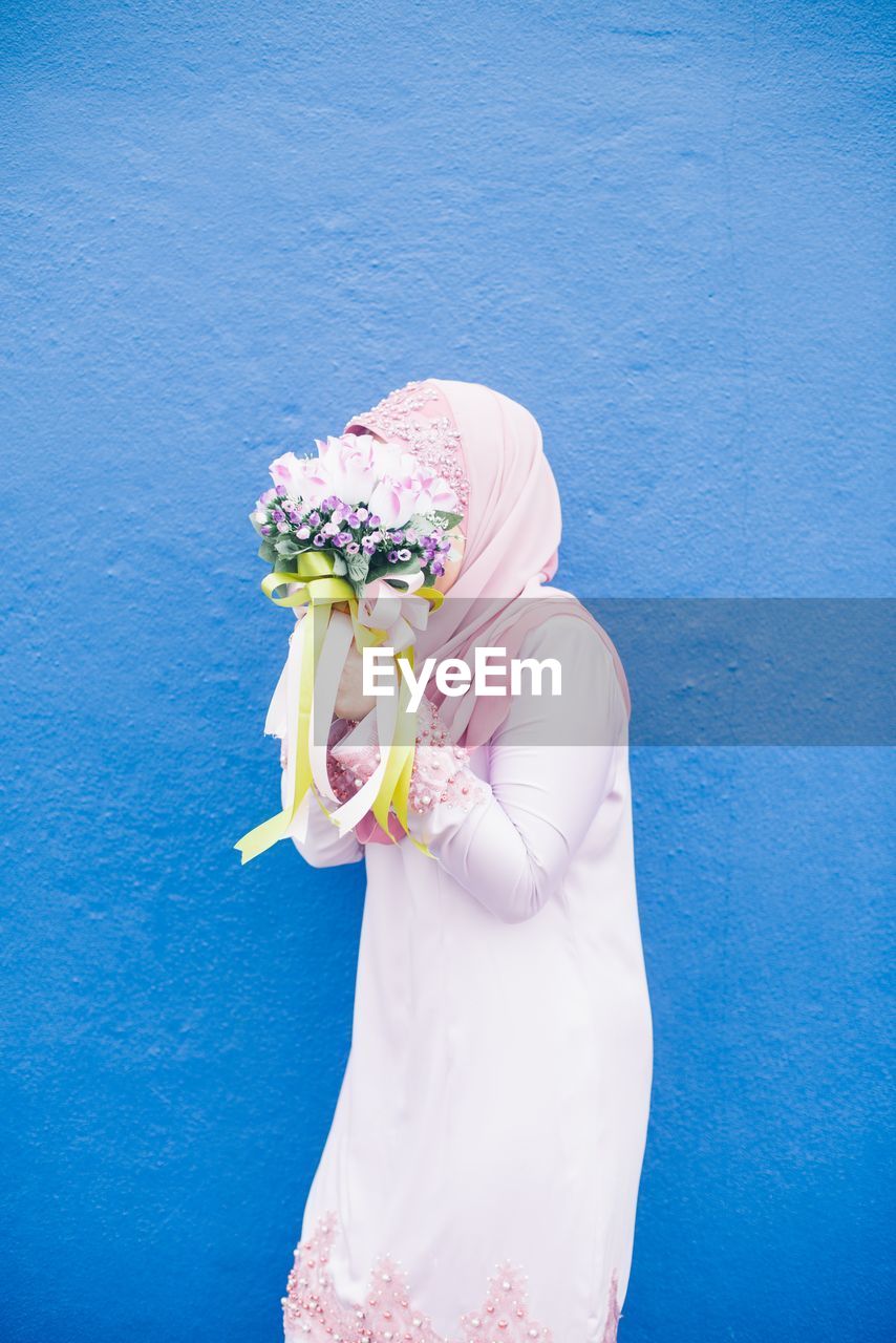 MIDSECTION OF WOMAN HOLDING BOUQUET AGAINST BLUE WALL