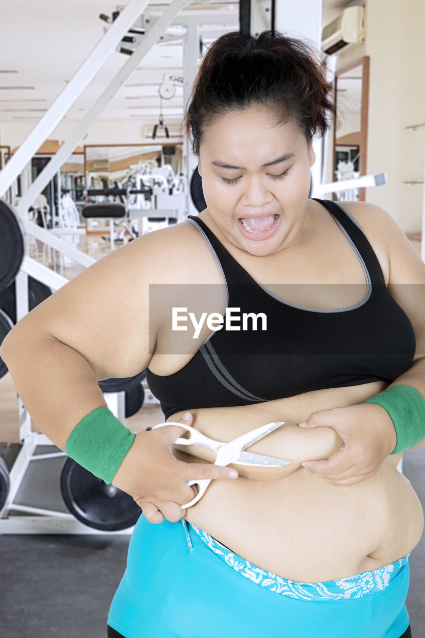 Overweight woman cutting abdomen with scissors in gym