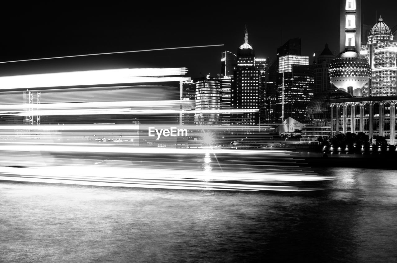 Light trails over river against buildings at night