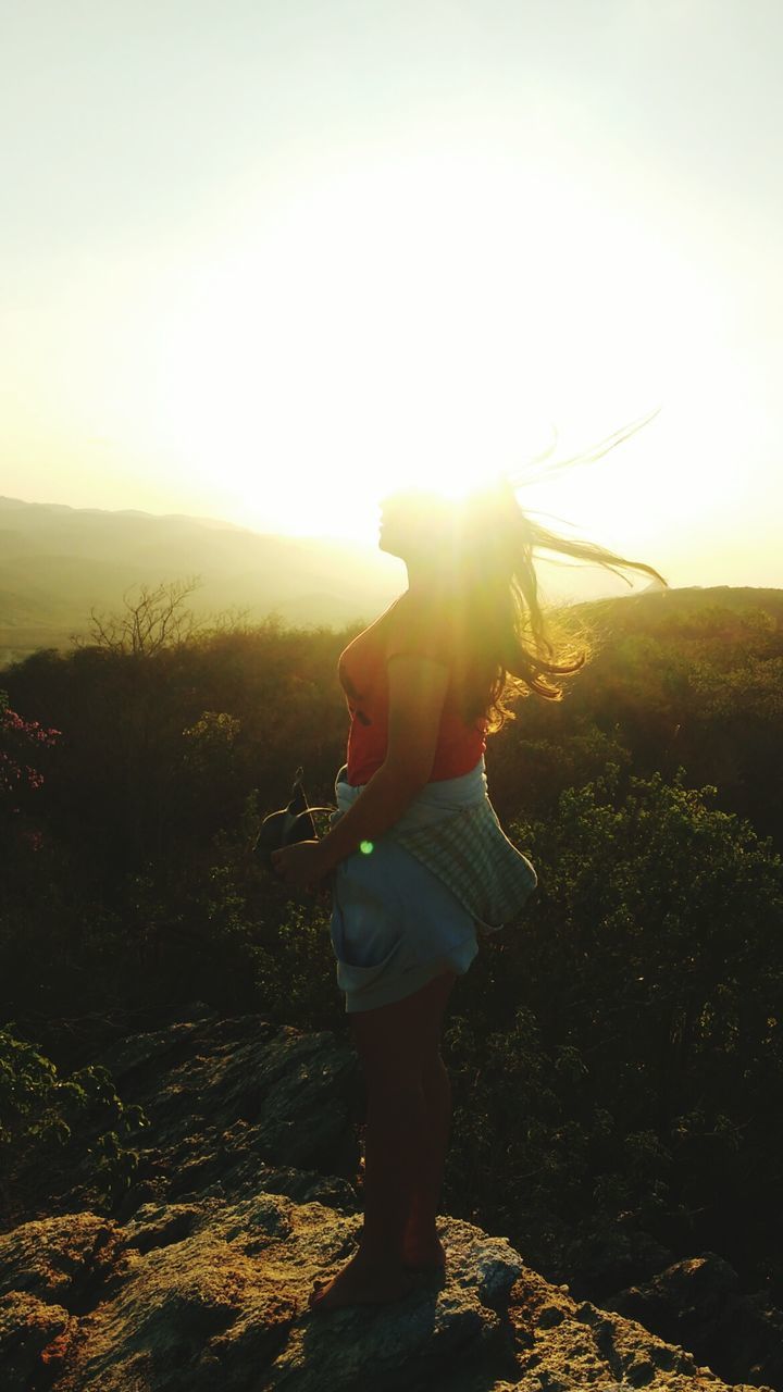 REAR VIEW OF WOMAN STANDING AGAINST SUNSET SKY