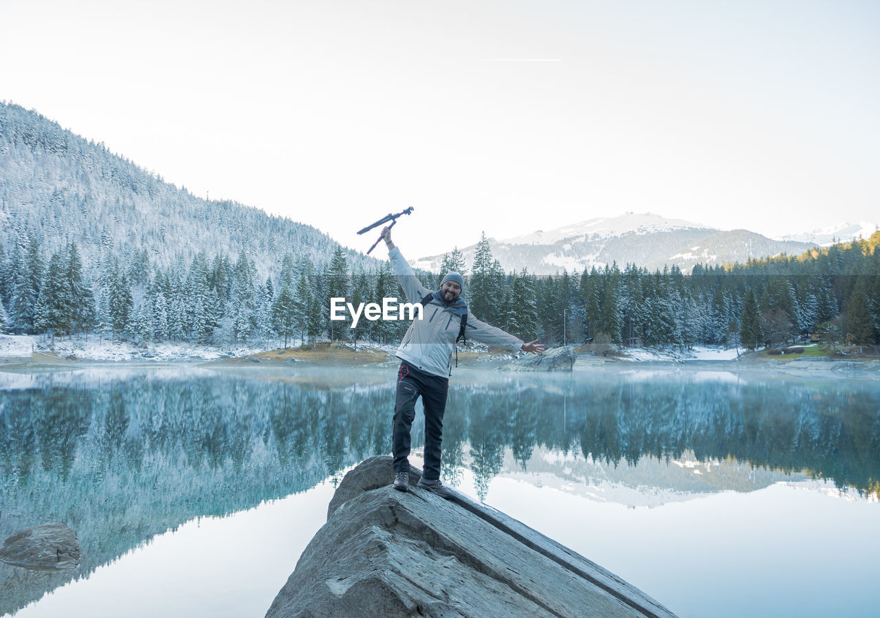 Portrait of smiling man standing with tripod on cliff against lake 