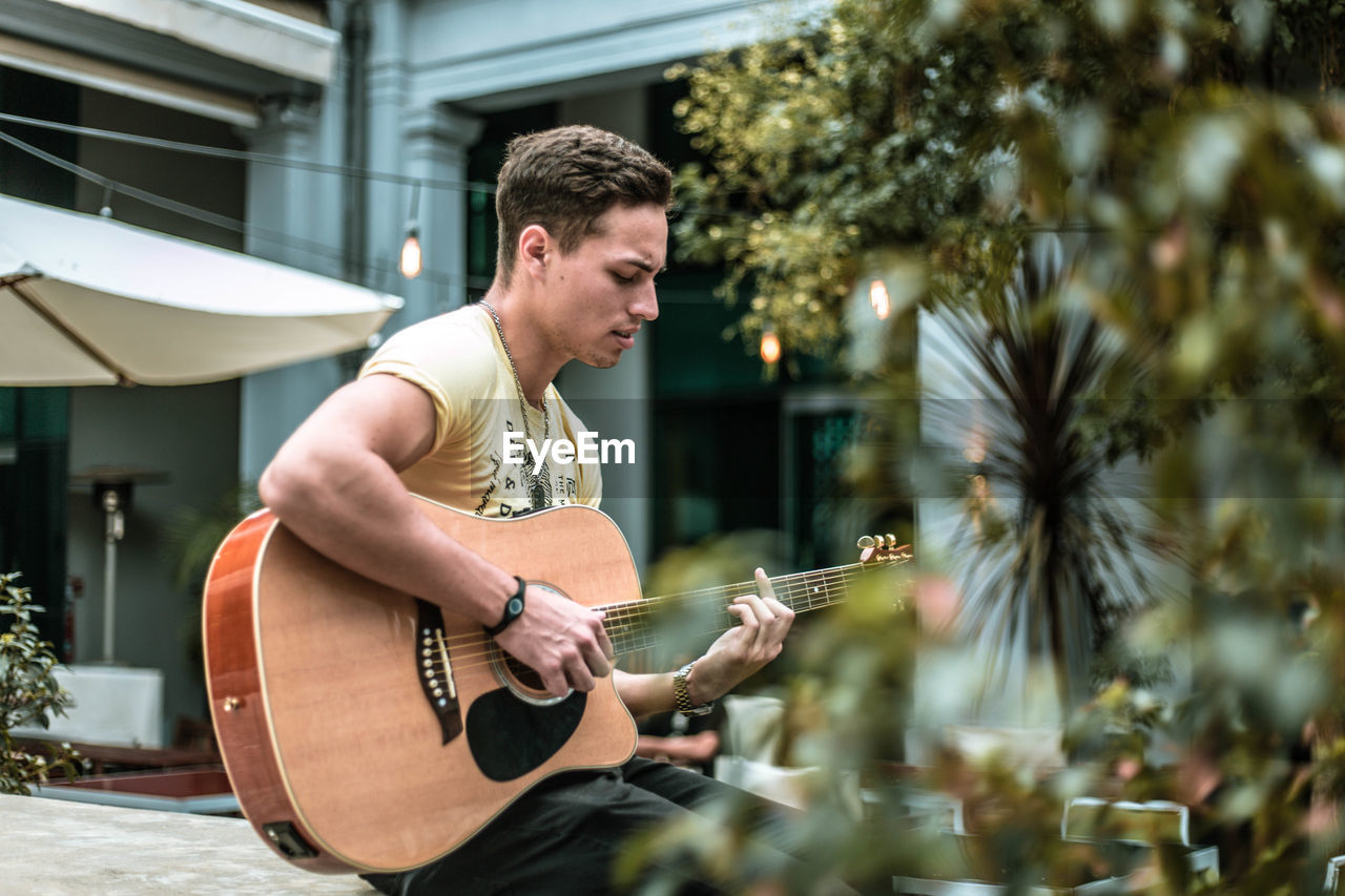 Young man playing guitar while sitting against building