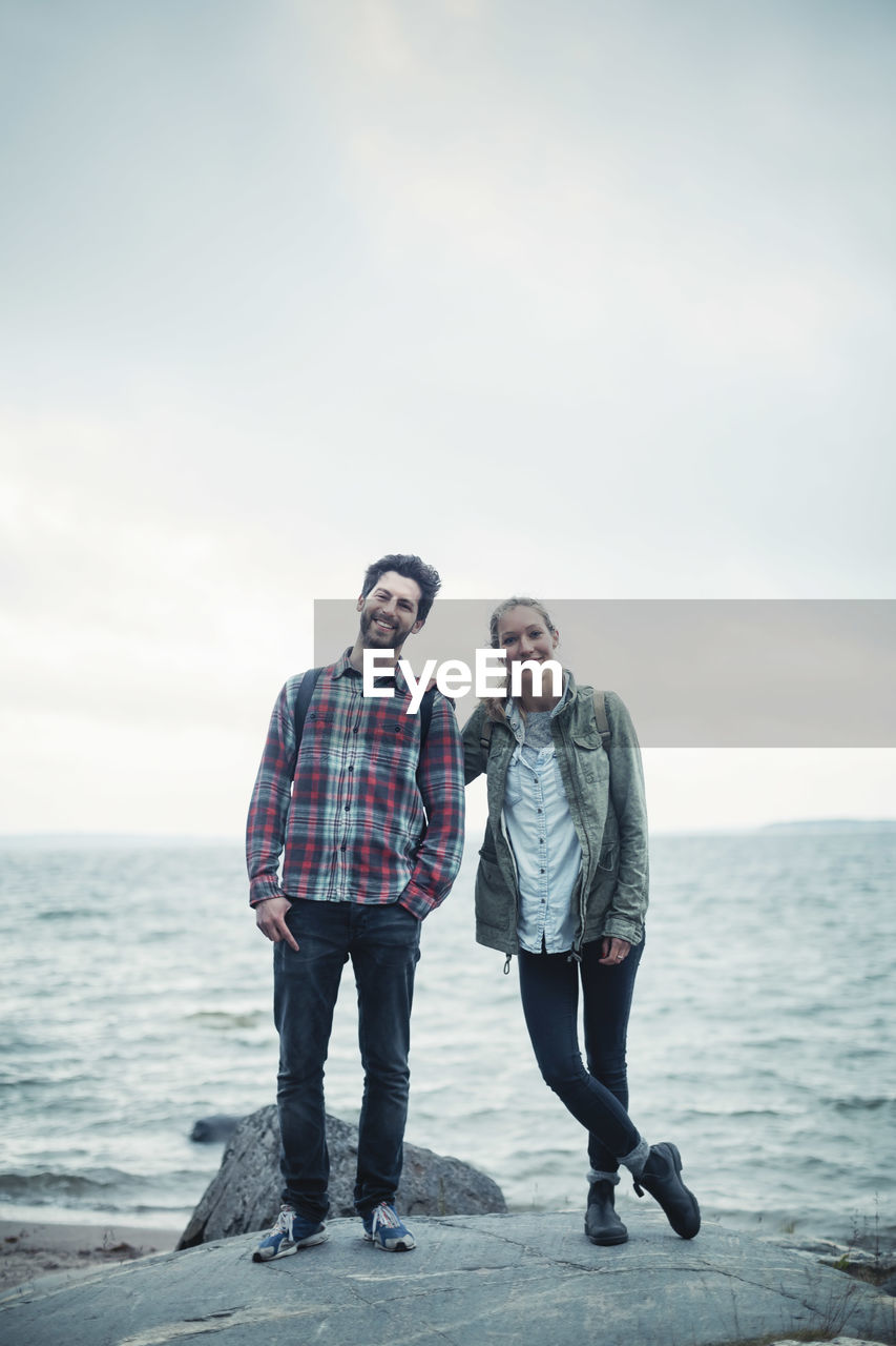 Full length portrait of confident wonderlust couple standing on rock by sea