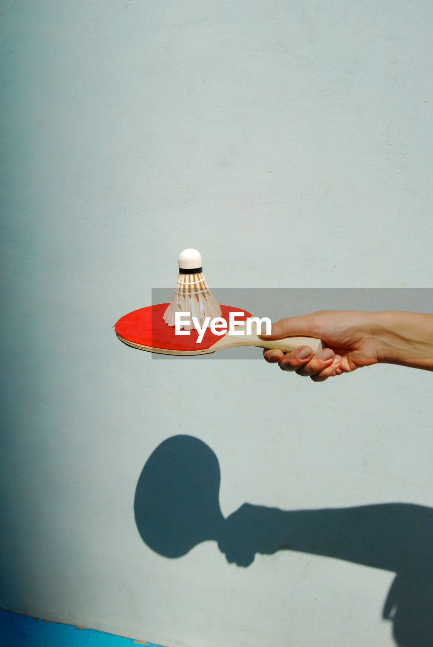 Close-up of human hand holding shuttle cock on tennis racket against wall