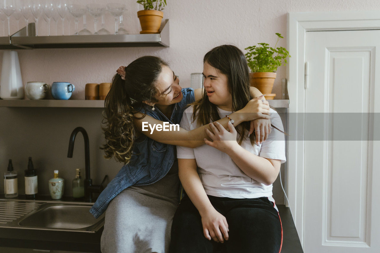 Smiling young woman hugging sister with down syndrome in kitchen at home