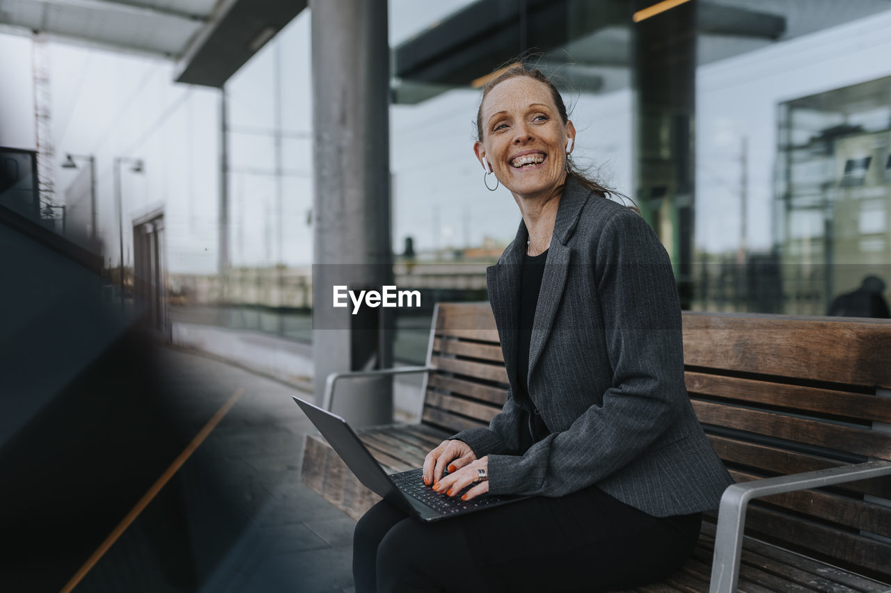 Cheerful businesswoman sitting with laptop on bench while waiting at railroad station