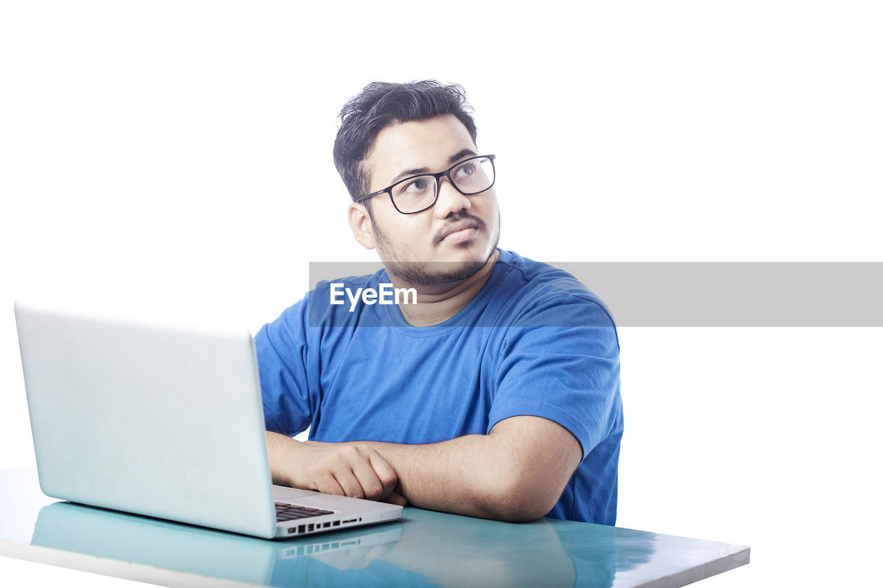 technology, computer, adult, eyeglasses, one person, laptop, wireless technology, men, using laptop, glasses, communication, business, casual clothing, sitting, indoors, internet, looking, person, table, businessman, writing, computer network, copy space, front view, working, portrait, office, conversation, white background, business finance and industry, furniture, desk, e-mail, beard, typing, relaxation, concentration, blue, young adult, occupation, facial hair, computer equipment, studio shot, lifestyles, new business