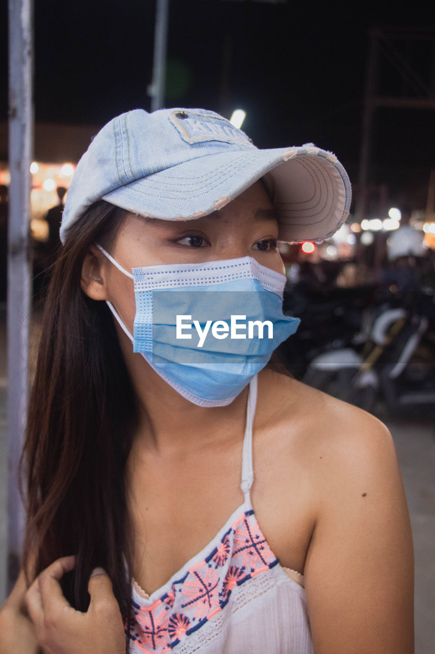 Close-up of young woman wearing flu mask and cap looking away standing outdoors