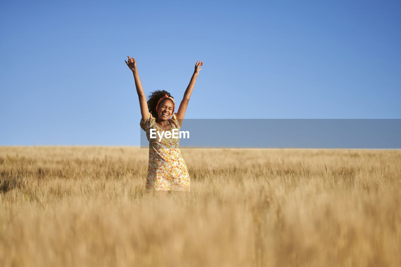Cheerful young woman with raised arms in wheat field standing in front of blue sky