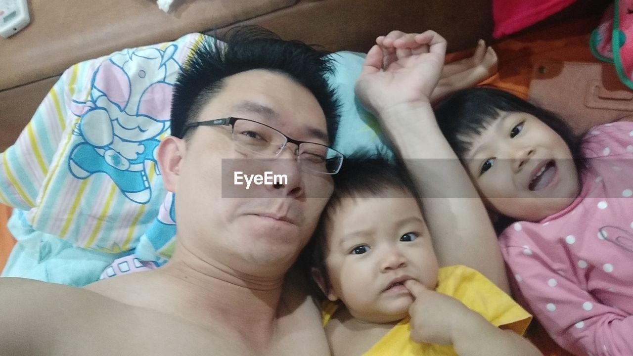 High angle portrait of shirtless father with children lying on bed at home