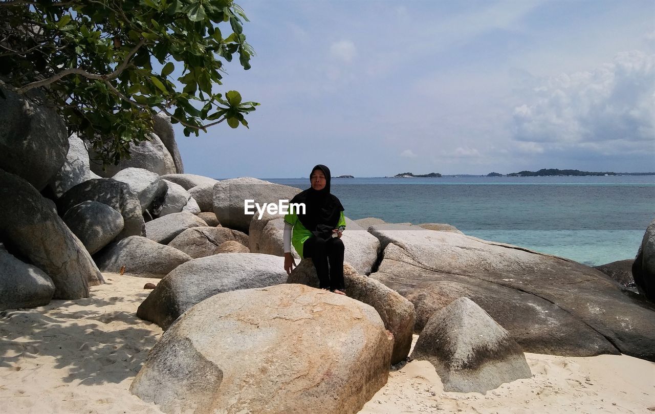 Man sitting on rock by sea against sky