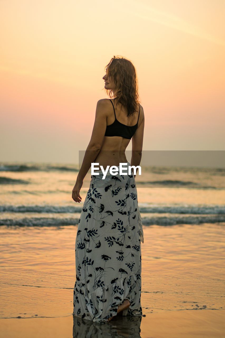 Woman standing at beach against sky during sunset