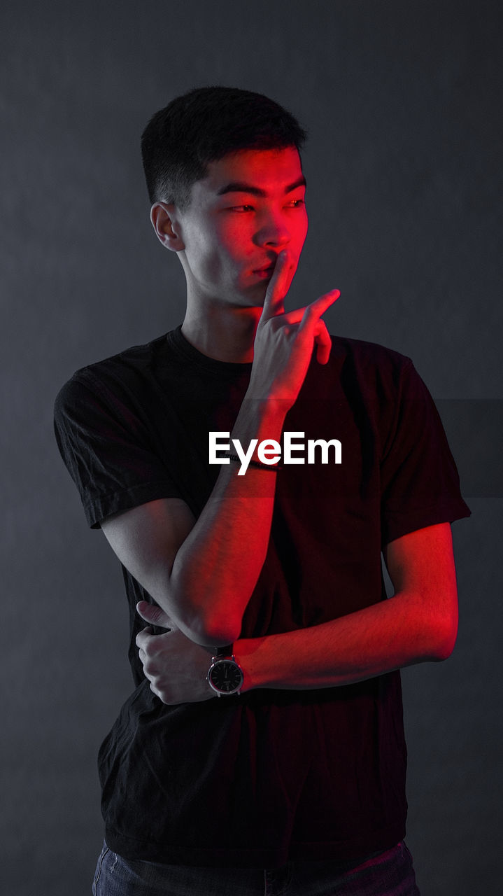 Young man with finger on lips looking away while standing against black background