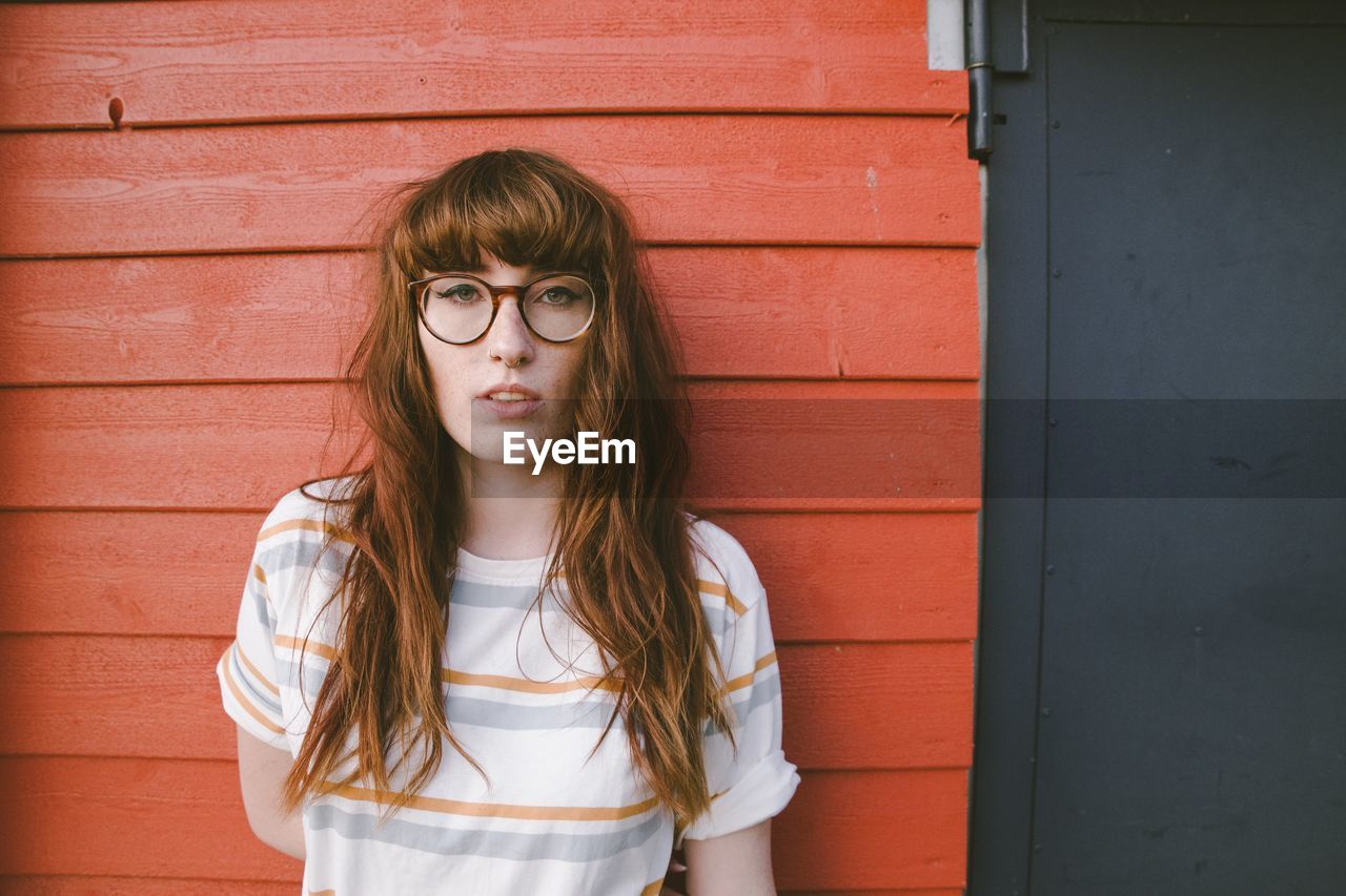PORTRAIT OF YOUNG WOMAN WEARING EYEGLASSES AGAINST WALL