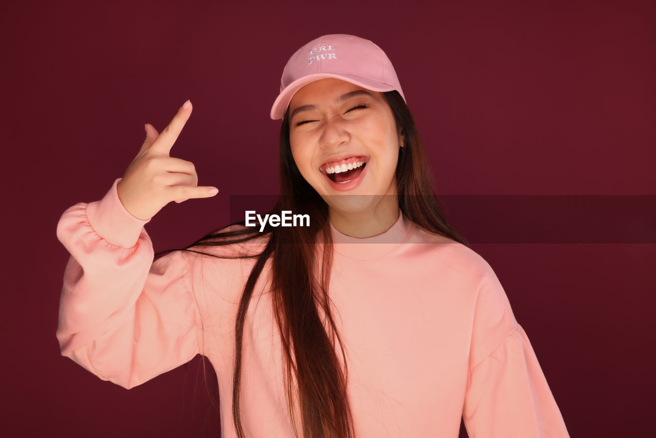 Portrait of happy young asian woman in the studio wearing pink clothes over garnet background. she is winking and making the rock sign