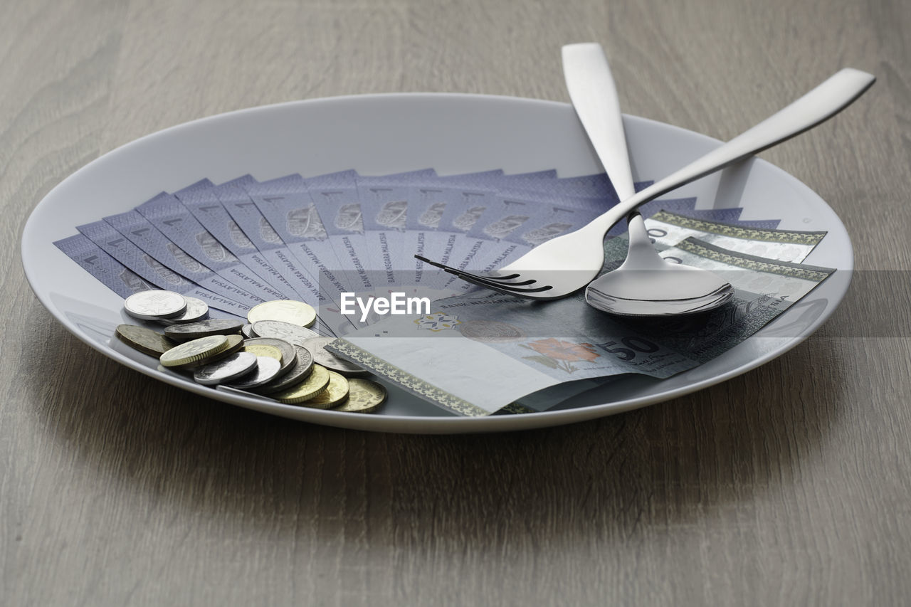 High angle view of paper currencies with coins and cutleries in plate on table