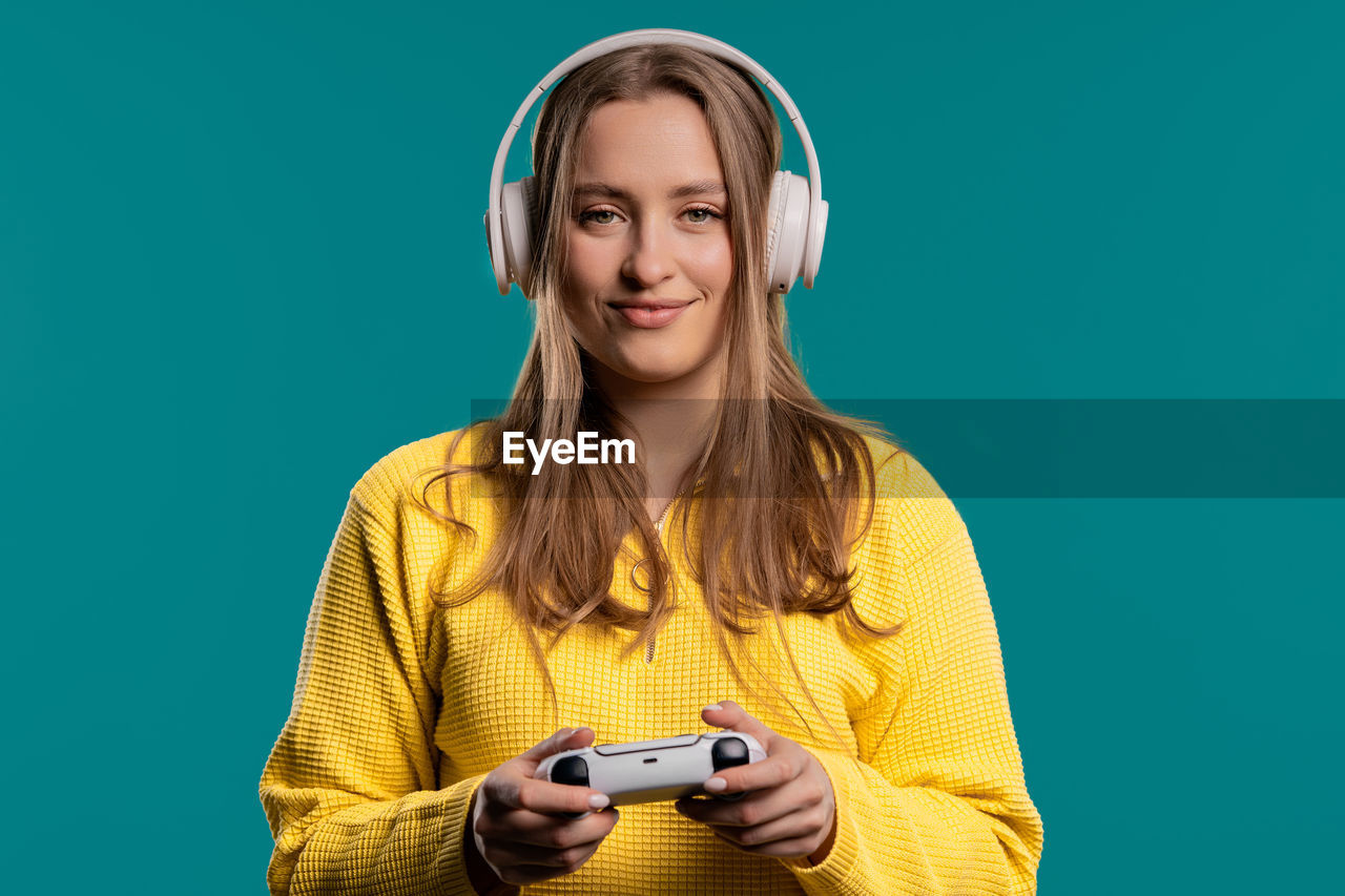 smiling, colored background, happiness, portrait, one person, technology, studio shot, women, emotion, adult, young adult, headphones, indoors, holding, music, long hair, fun, cheerful, listening, casual clothing, arts culture and entertainment, looking at camera, person, waist up, hairstyle, enjoyment, communication, front view, yellow, child, blond hair, human face, copy space, wireless technology, positive emotion, blue, clothing, retro styled, lifestyles, leisure activity, teenager, standing