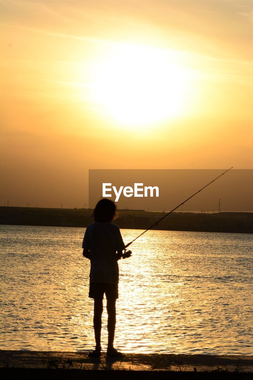 Rear view of silhouette man fishing in sea against sunset sky