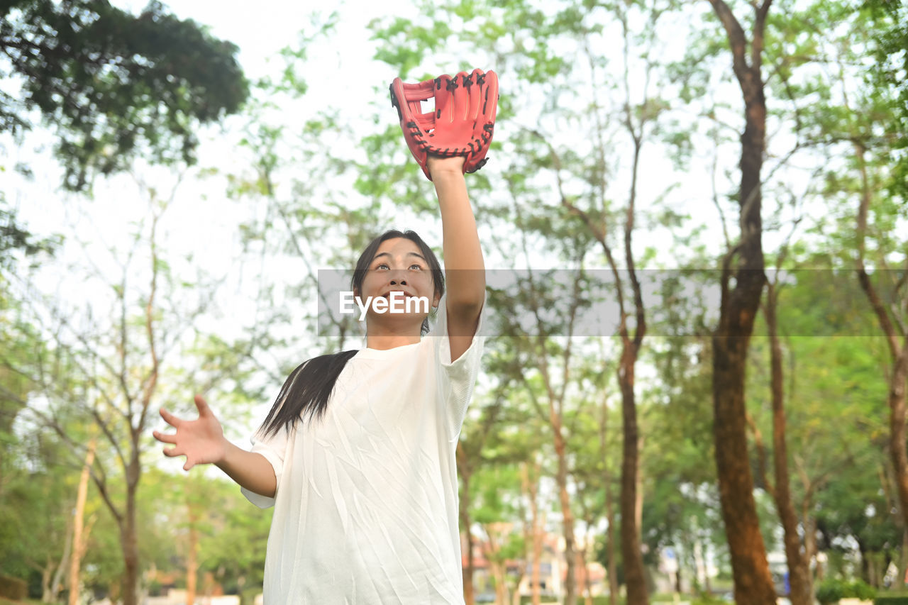 portrait of smiling young woman with arms raised standing against trees