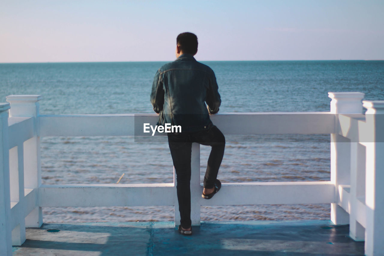 REAR VIEW OF MAN STANDING ON SEA SHORE AGAINST SKY