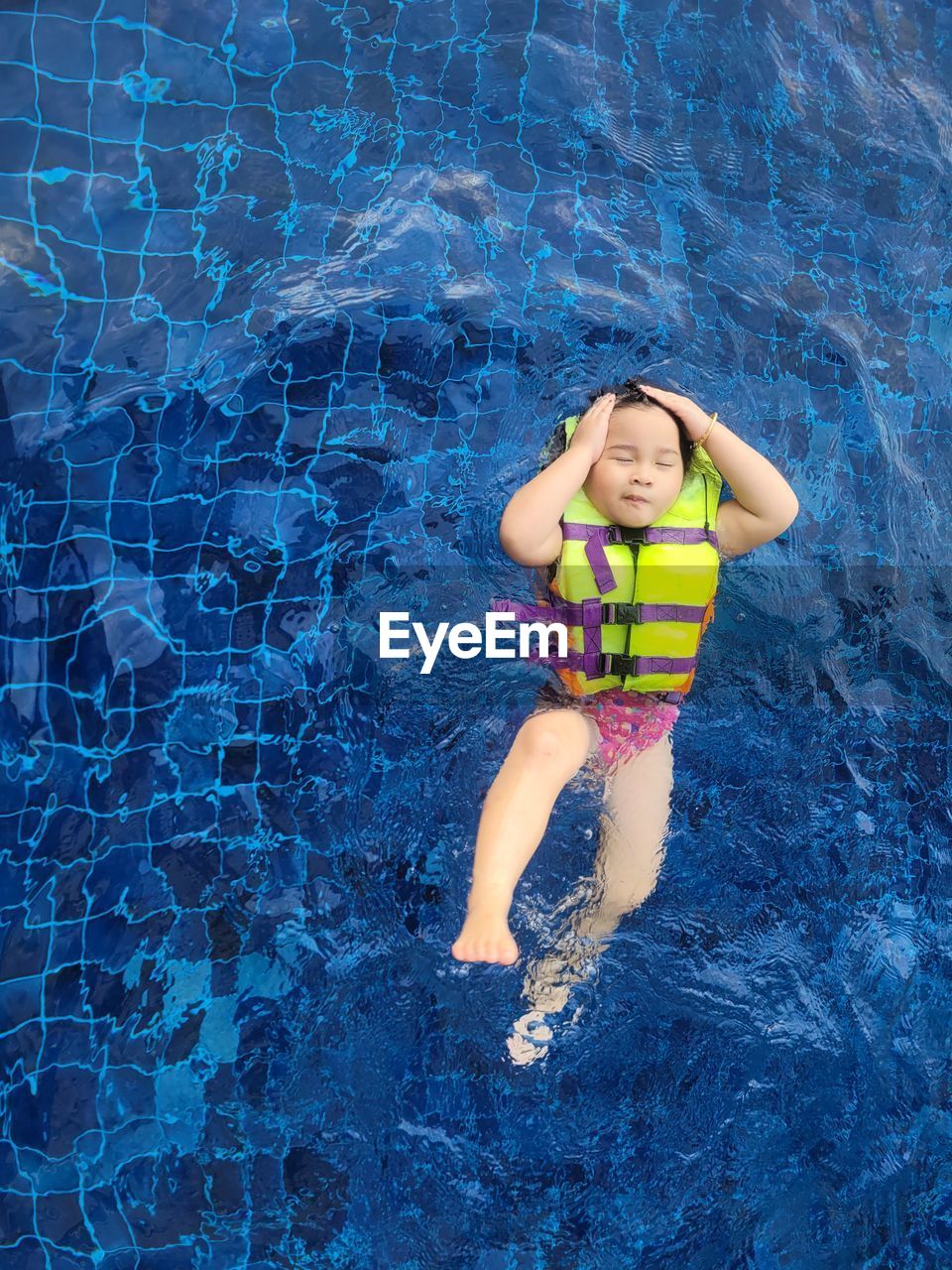 A cute asian girl wearing a pink swimsuit and a green life jacket is playing in the swimming pool.