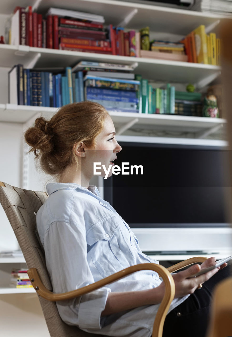 Side view of woman holding digital tablet while sitting on chair at home