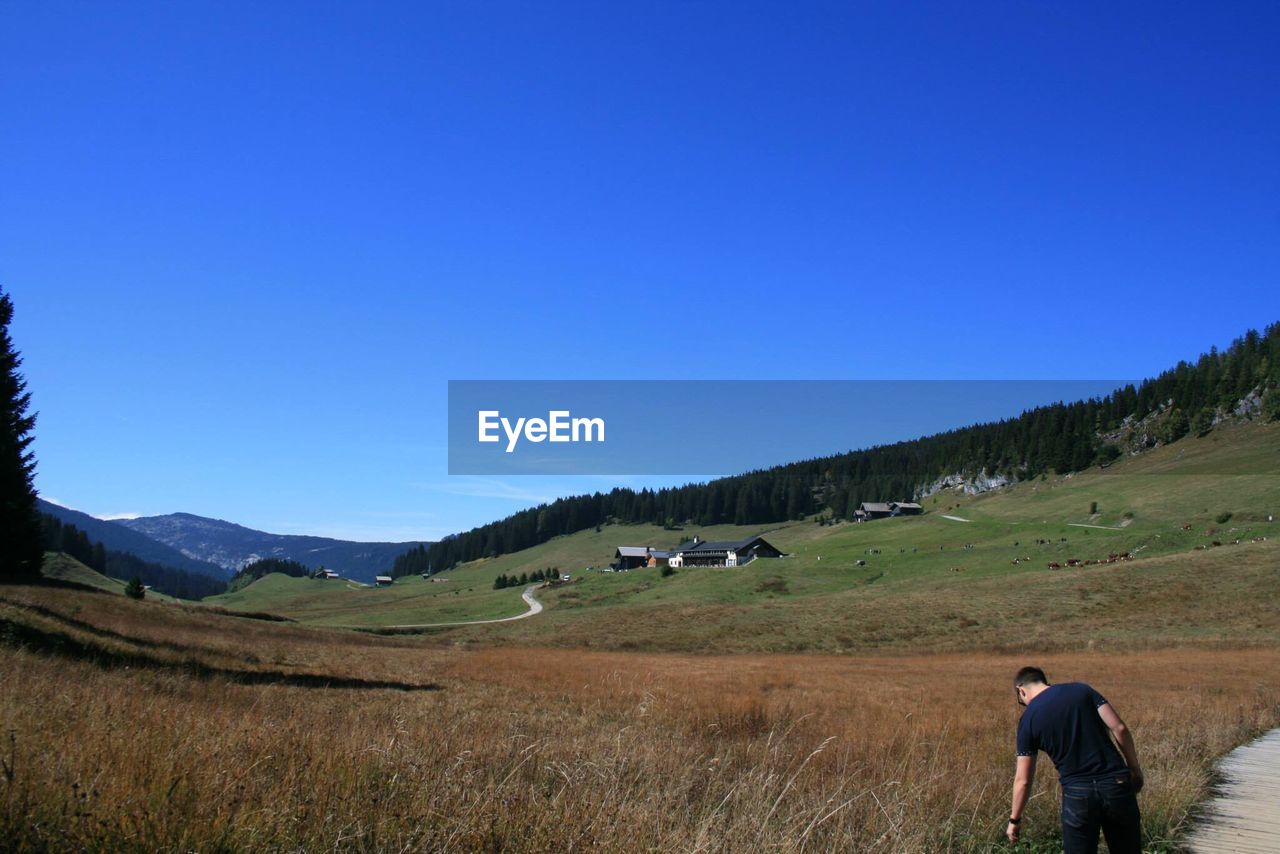 REAR VIEW OF MAN STANDING ON FIELD AGAINST MOUNTAIN