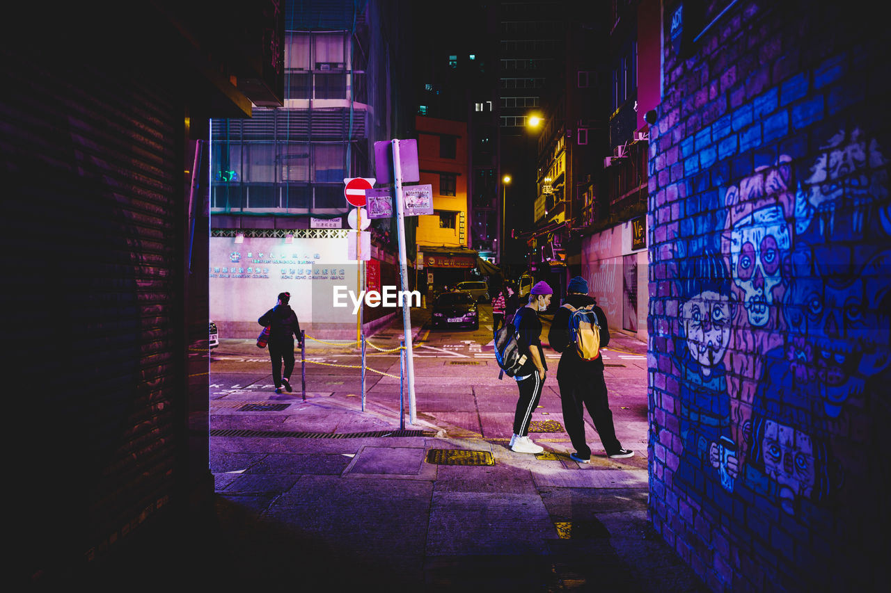 PEOPLE WALKING ON ILLUMINATED STREET AMIDST BUILDINGS