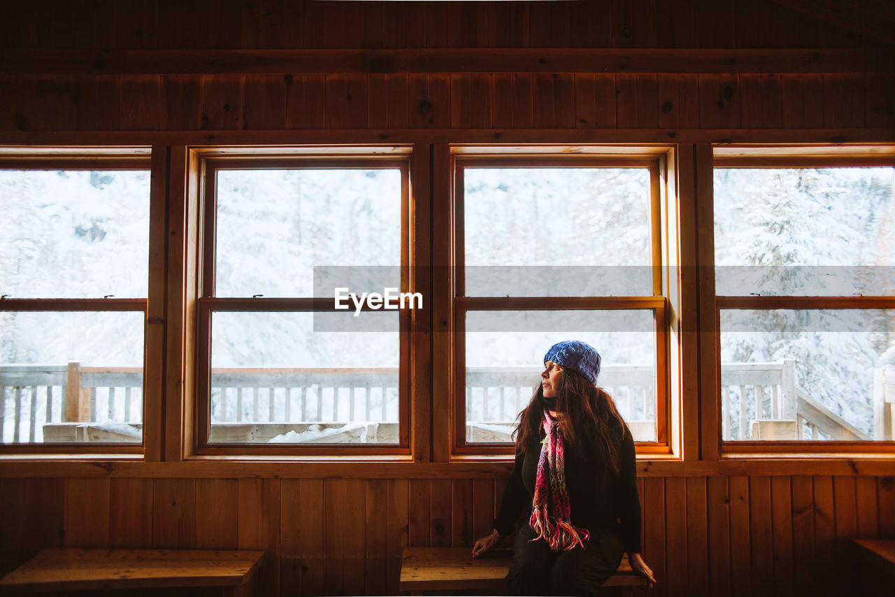 Woman in outerwear sitting on bench near windows and looking away inside lumber hut on winter day in valley of the ghosts in monts valin national park in quebec, canada