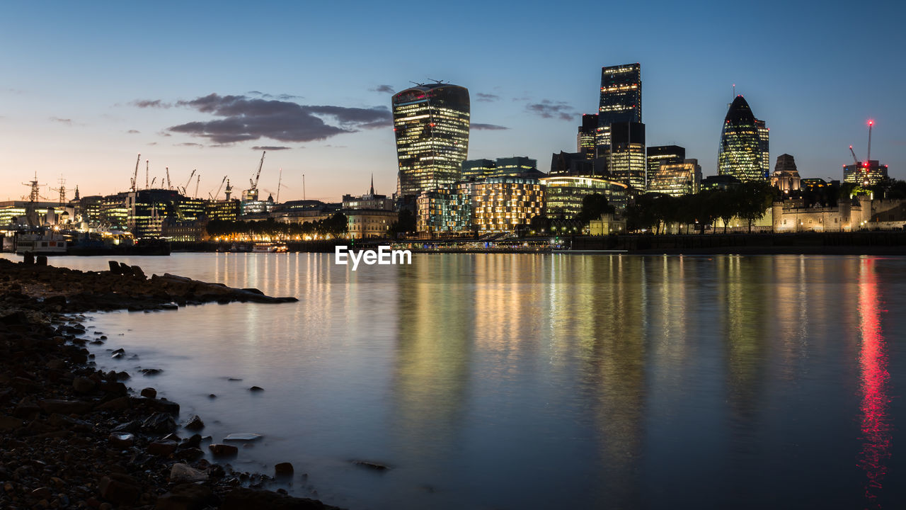 Illuminated modern buildings by thames river against sky in city at dusk