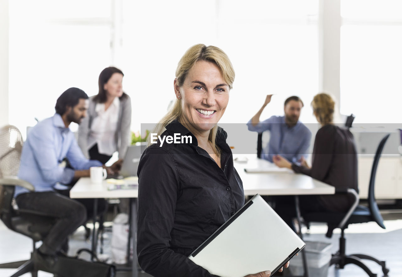 Portrait of mature businesswoman holding laptop with colleagues in background at office