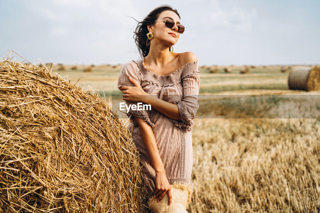 Smiling woman in sunglasses with bare shoulders on a background of wheat field and bales of hay.