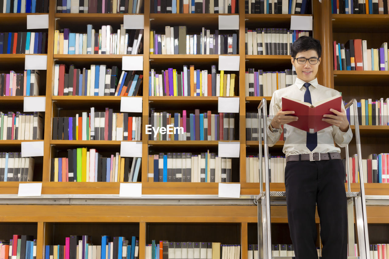 Smiling wearing man wearing eyeglasses reading book in library