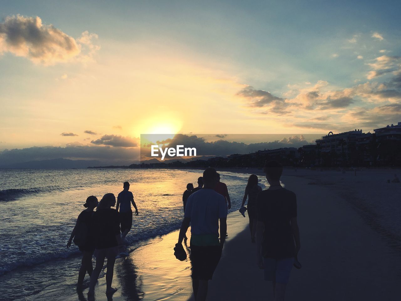 REAR VIEW OF PEOPLE ENJOYING AT BEACH DURING SUNSET