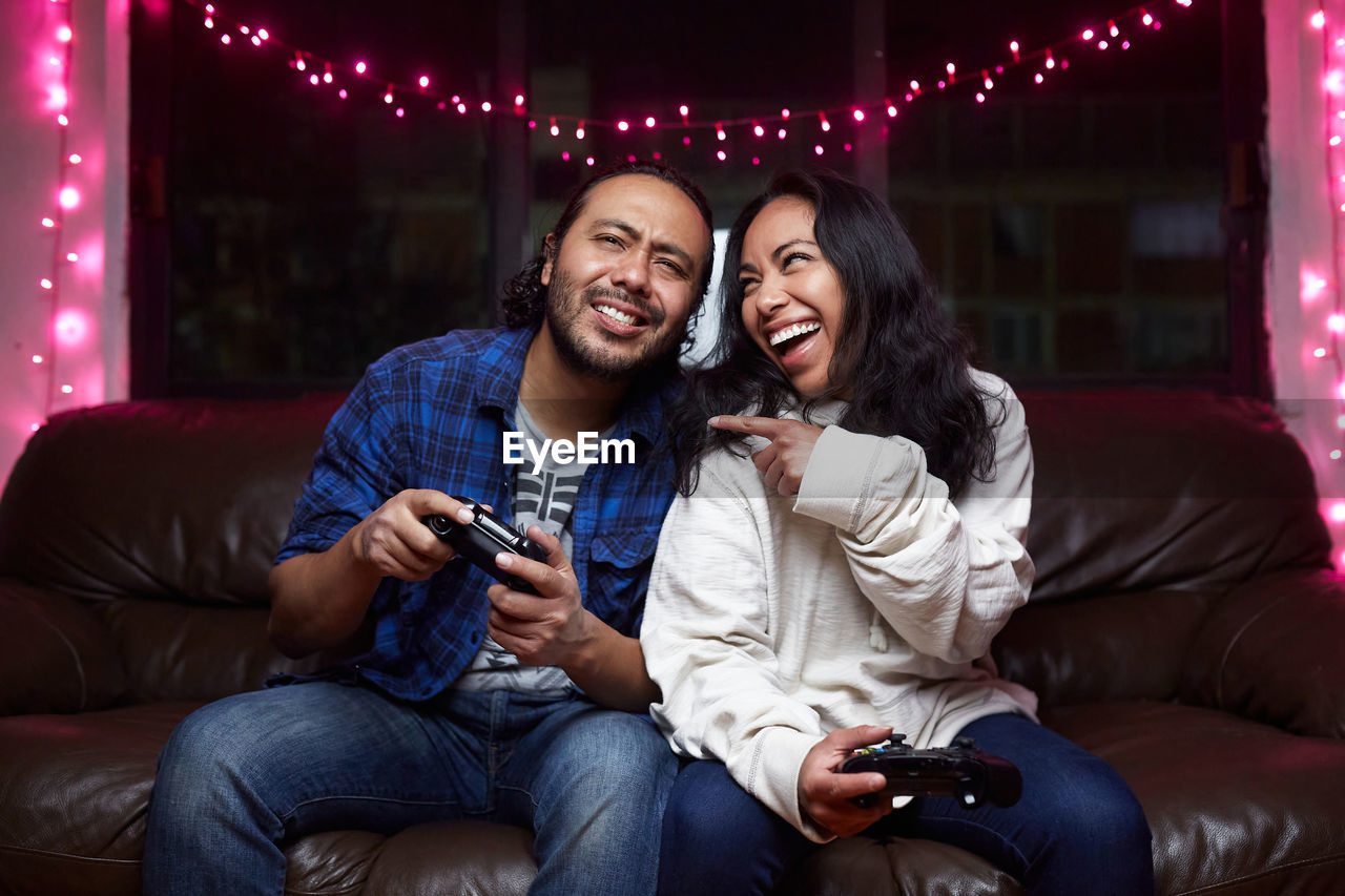 Excited ethnic couple in casual wear with joy pads playing video game together while sitting on leather couch at home
