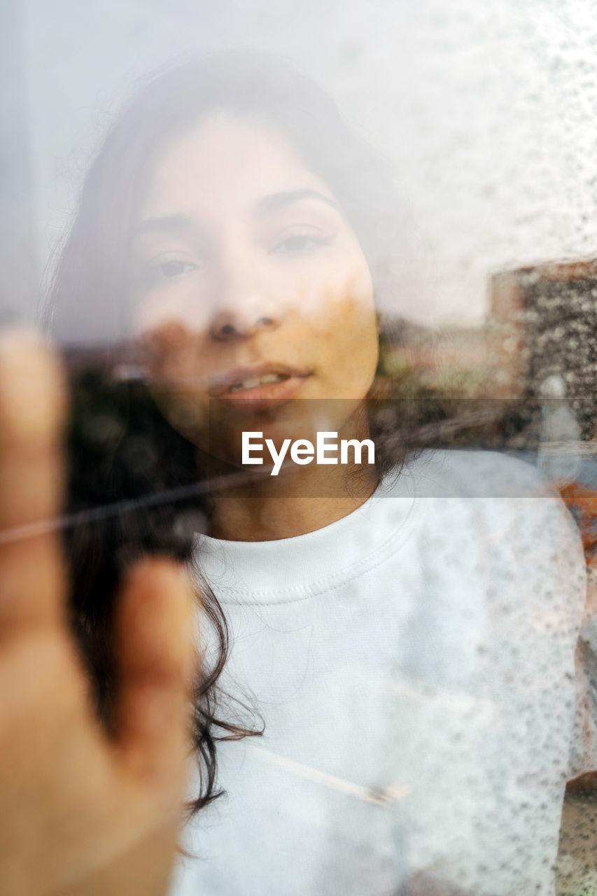 Through glass of young ethnic female gently touching window with raindrops and looking at camera